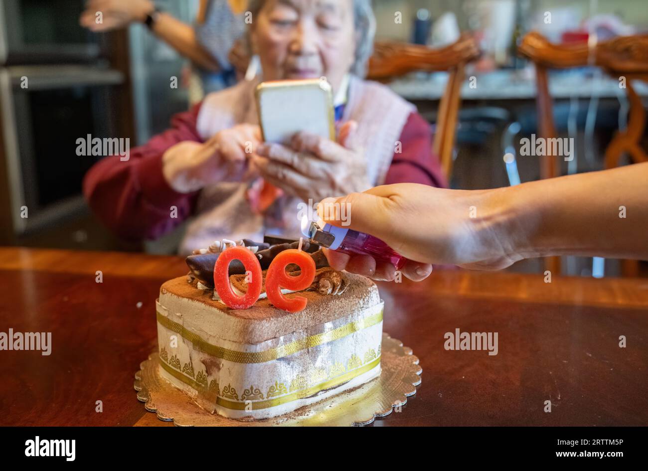 Donna anziana che scatta foto da smartphone di una torta che festeggia il suo 90° compleanno. Le mani del nipote illuminano le candele. Foto Stock