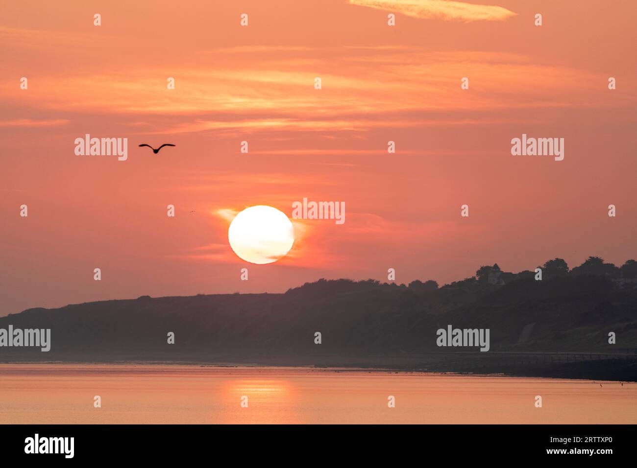 Il sorgere del sole sulla costa distanti sagomata di Reculver nel Kent, e in primo piano il mare calmo che riflette il colore arancione del cielo, alcuni sottili strati di nuvole. Foto Stock