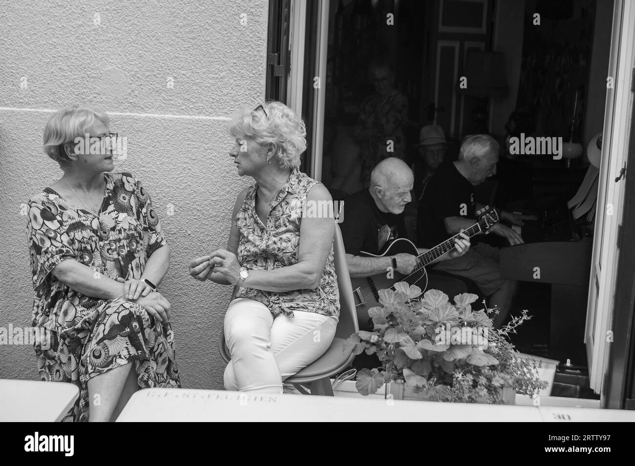 Due donne che parlano mentre gli uomini suonano musica nella stanza dietro, Lachaux, Saint-Felicien, Ardeche, Francia Foto Stock