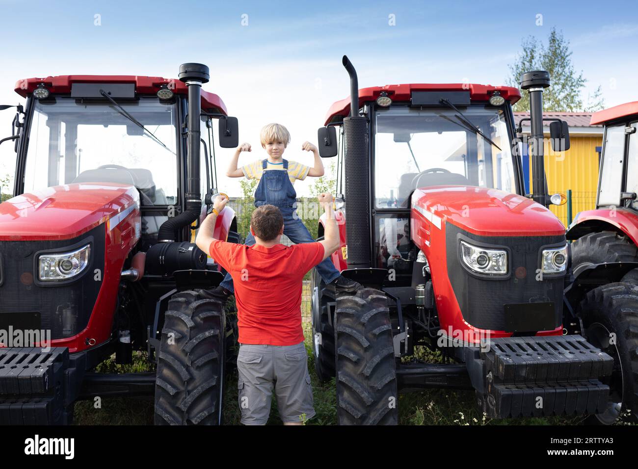 uomo e ragazzo si trovano di fronte ai nuovi trattori rossi di grandi dimensioni presso il sito di vendita. Un adulto e un bambino guardano con interesse le macchine agricole e mostrano il loro potere Foto Stock