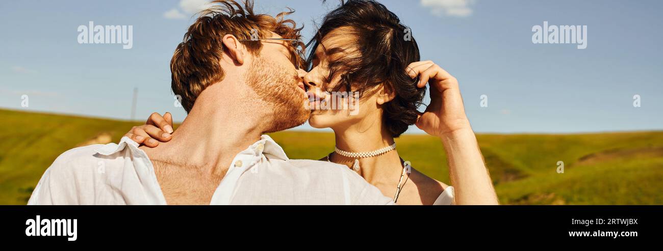 matrimonio rustico, giovani sposi interrazziali che si baciano sul vento sotto il cielo blu in campo verde, banner Foto Stock