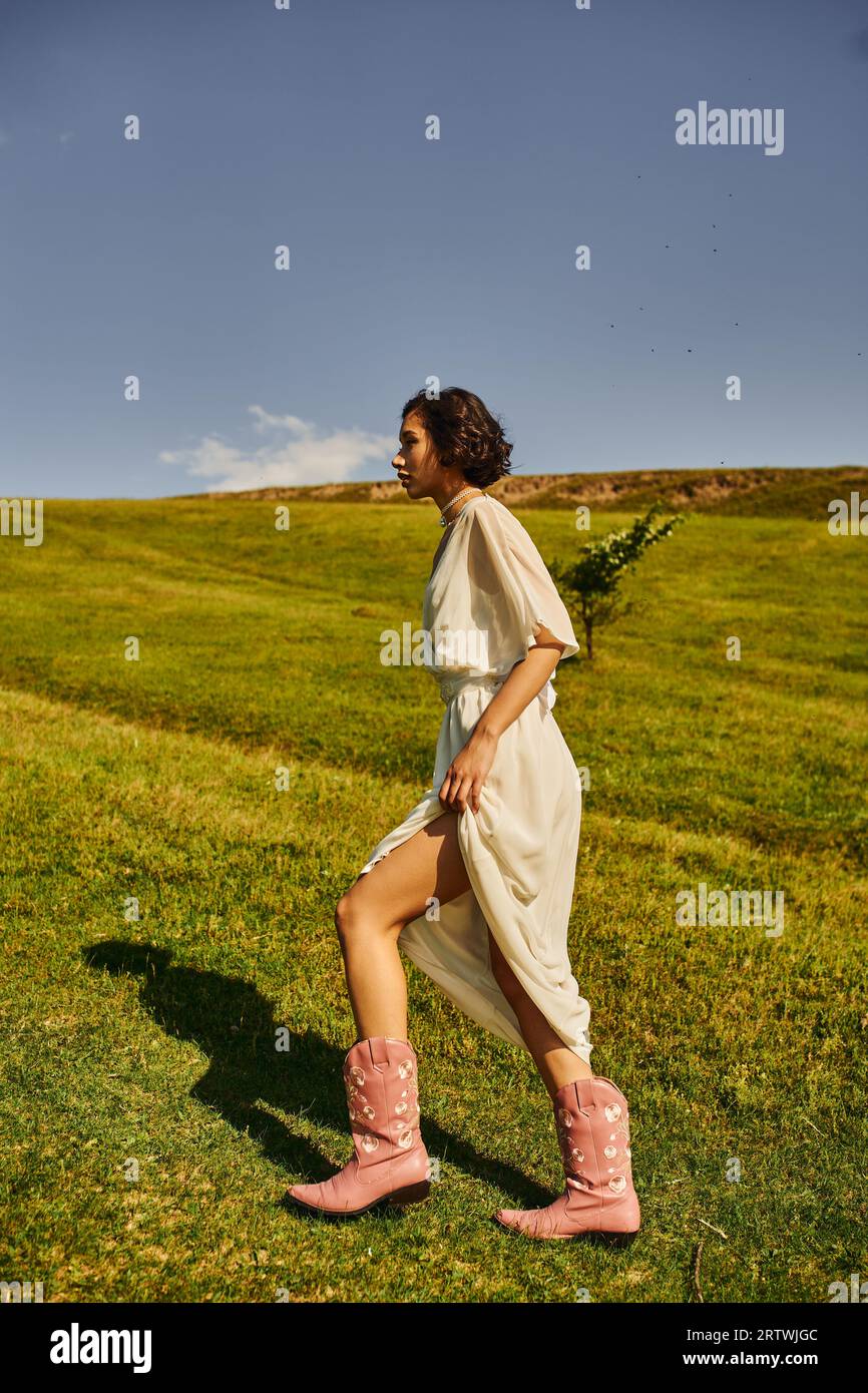 vista laterale della giovane donna asiatica in abito da sposa e stivali da cowboy che camminano sul prato in campagna Foto Stock