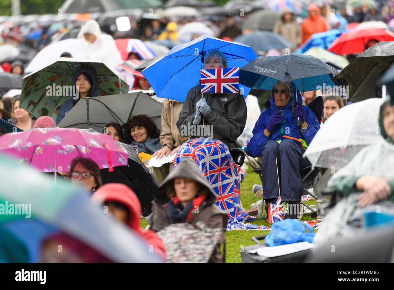 Una donna al riparo dalla pioggia sotto un ombrello ha una bandiera Union Jack. Fa parte di una grande folla di persone che assistono all'incoronazione di re CH Foto Stock