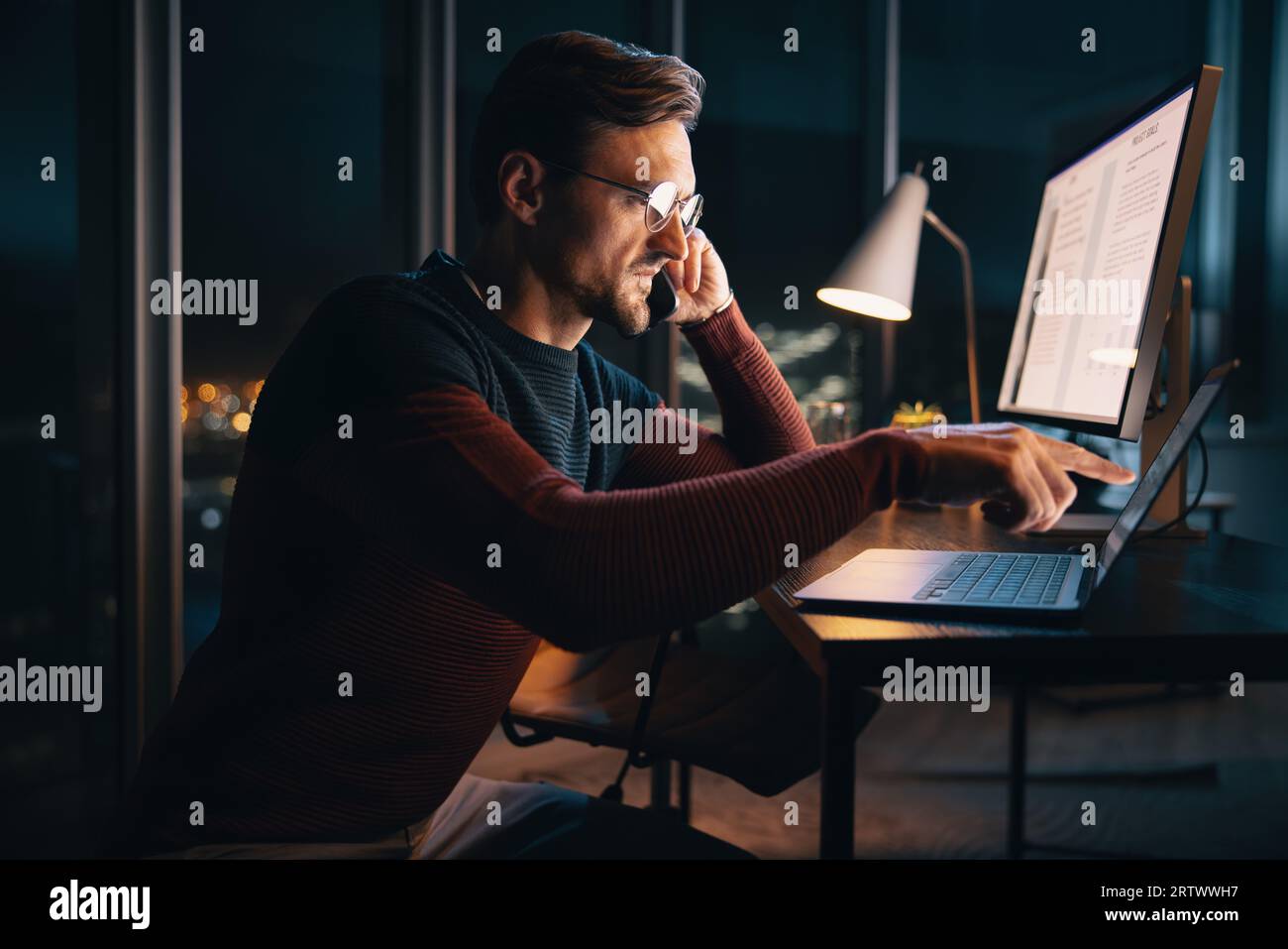 Un uomo d'affari caucasico lavora a tarda notte in un ufficio, discutendo di un progetto al telefono. È concentrato e determinato, bilanciando la tecnologia e. Foto Stock