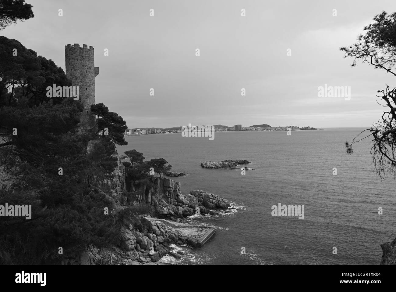 Una vista in scala di grigi di una collina rocciosa con una torre che si affaccia sull'acqua della Costa Brava Foto Stock