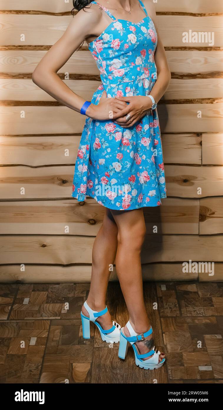 Giovane personaggio femminile sottile con le mani sulla cintura e sulle gambe in posa con un abito blu a motivi floreali e sandali al chiuso. Foto Stock