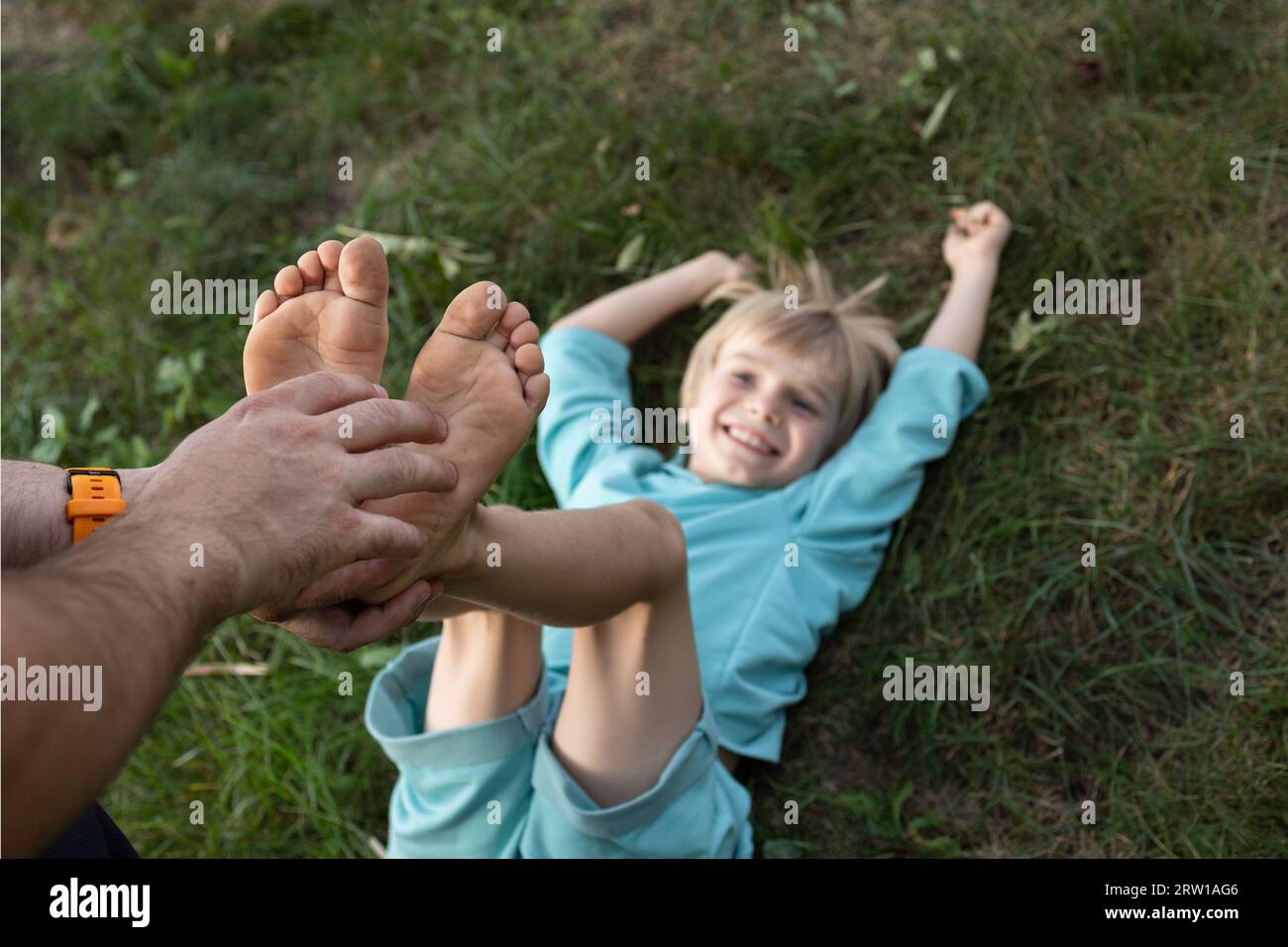 le mani dell'uomo solleticano i piedi nudi di di un bambino sdraiato sull'erba. Divertimento in famiglia, infanzia felice. giorno del figlio. La gioia della comunicazione, giocare ad aro Foto Stock