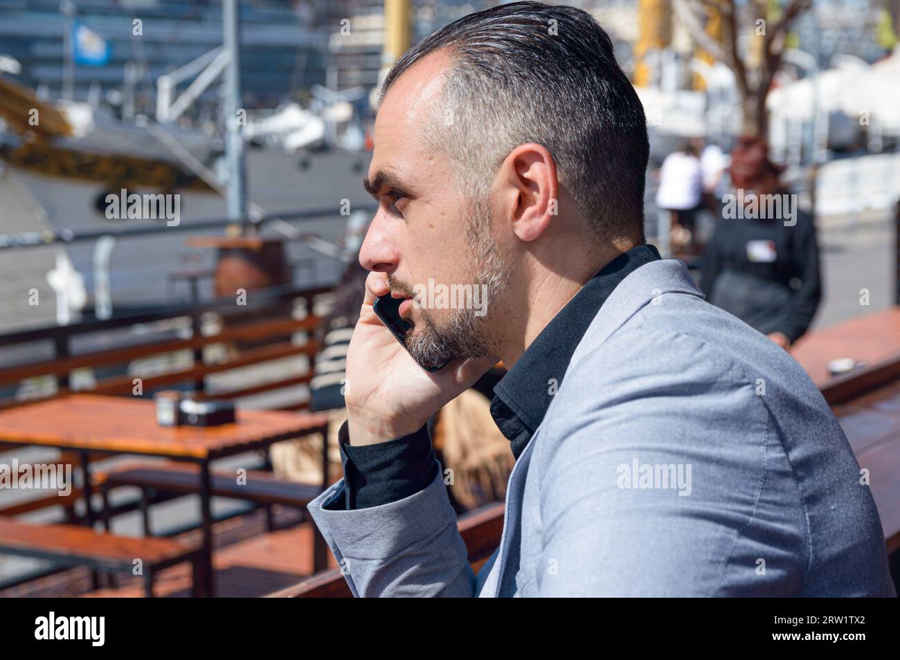 Vista del profilo di un elegante uomo d'affari caucasico con barba in giacca grigia seduto fuori dal ristorante mentre parla al telefono Foto Stock