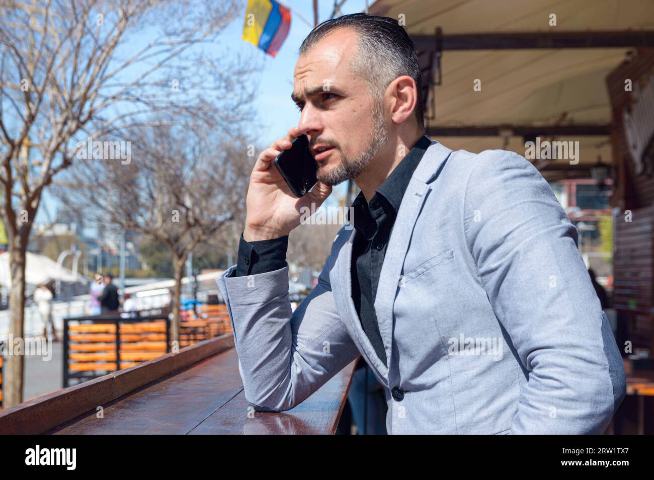 Vista del profilo di un elegante uomo d'affari caucasico con barba in giacca grigia seduto fuori dal ristorante mentre parla al telefono Foto Stock