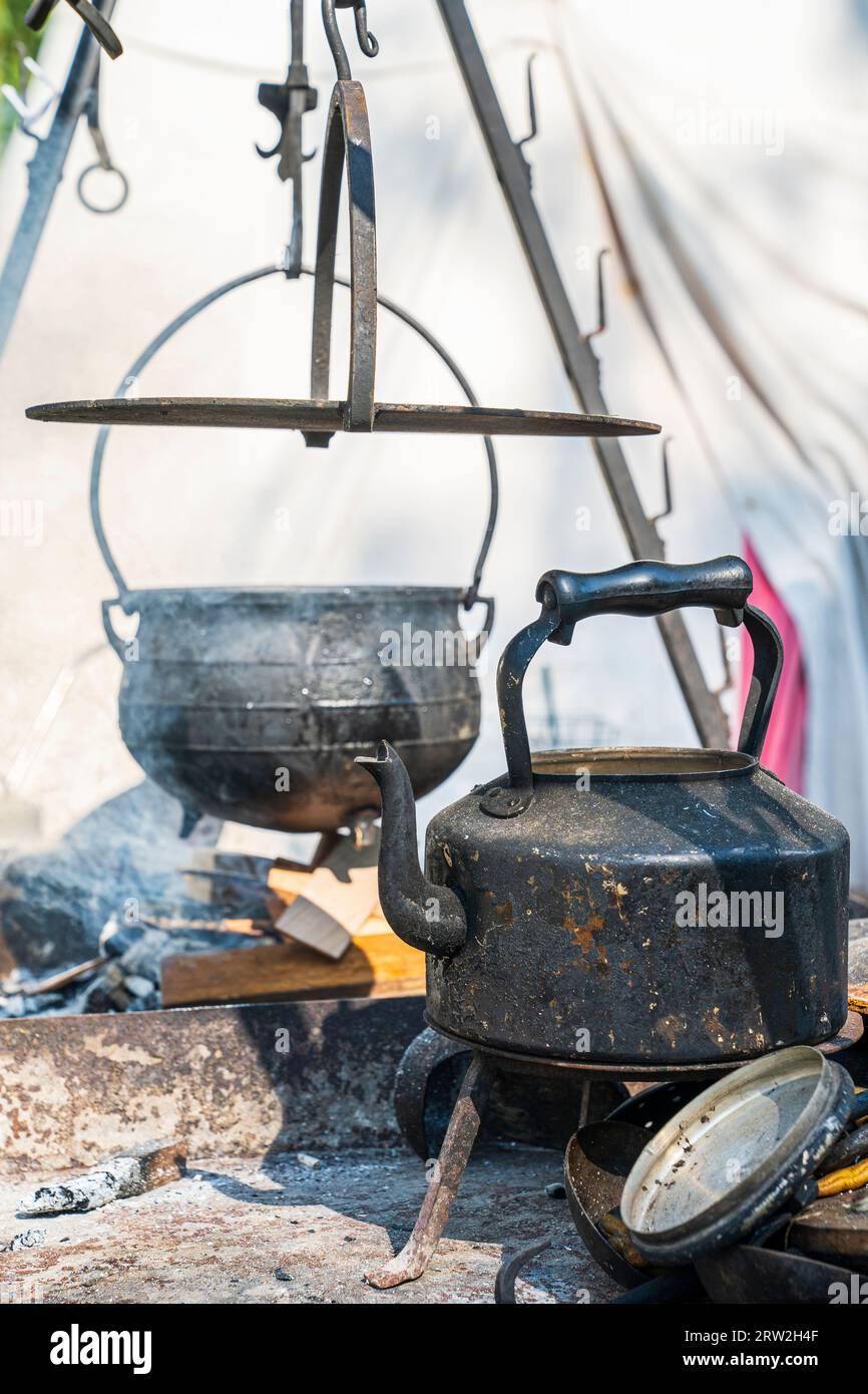 Bollitore nero in ferro medievale su un supporto in ghisa, dietro, un calderone sfocato appeso su un fuoco in fiamme. Centro sandwich medievale. Foto Stock