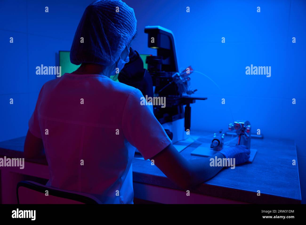 Giovane scienziato in maschera protettiva lavora con biomateriale sotto microscopio Foto Stock