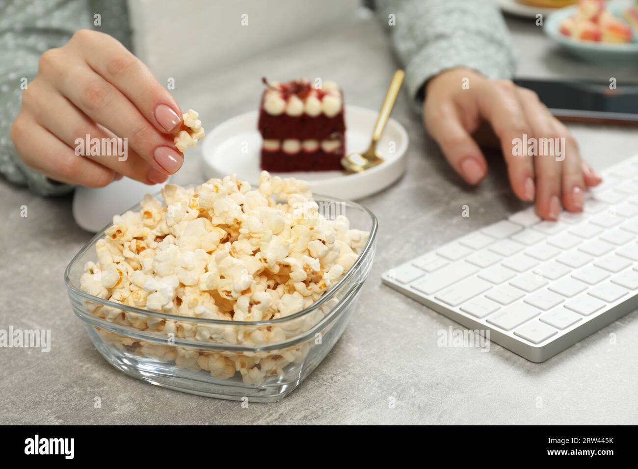 Cattive abitudini. Donna che mangia popcorn mentre lavora al computer al tavolo grigio chiaro, primo piano Foto Stock