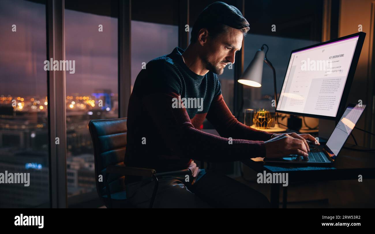 Uomo d'affari caucasico concentrato che digita il suo portatile a una scrivania in un ufficio notturno. Uomo professionista che lavora a un piano di marketing digitale, mostrando Foto Stock