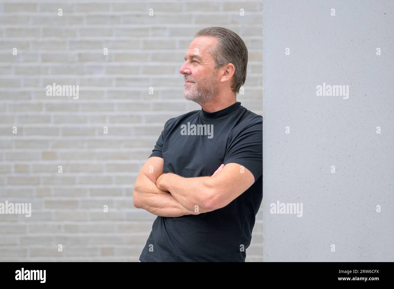 L'attraente uomo dai capelli grigi si appoggia contro un muro con le braccia incrociate e guarda in lontananza davanti a un muro di mattoni bianchi Foto Stock