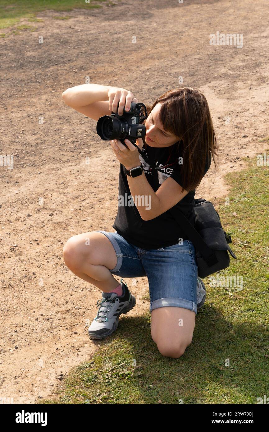 Giovane fotografa in ginocchio mentre scatta foto con la fotocamera DSLR all'aperto Foto Stock