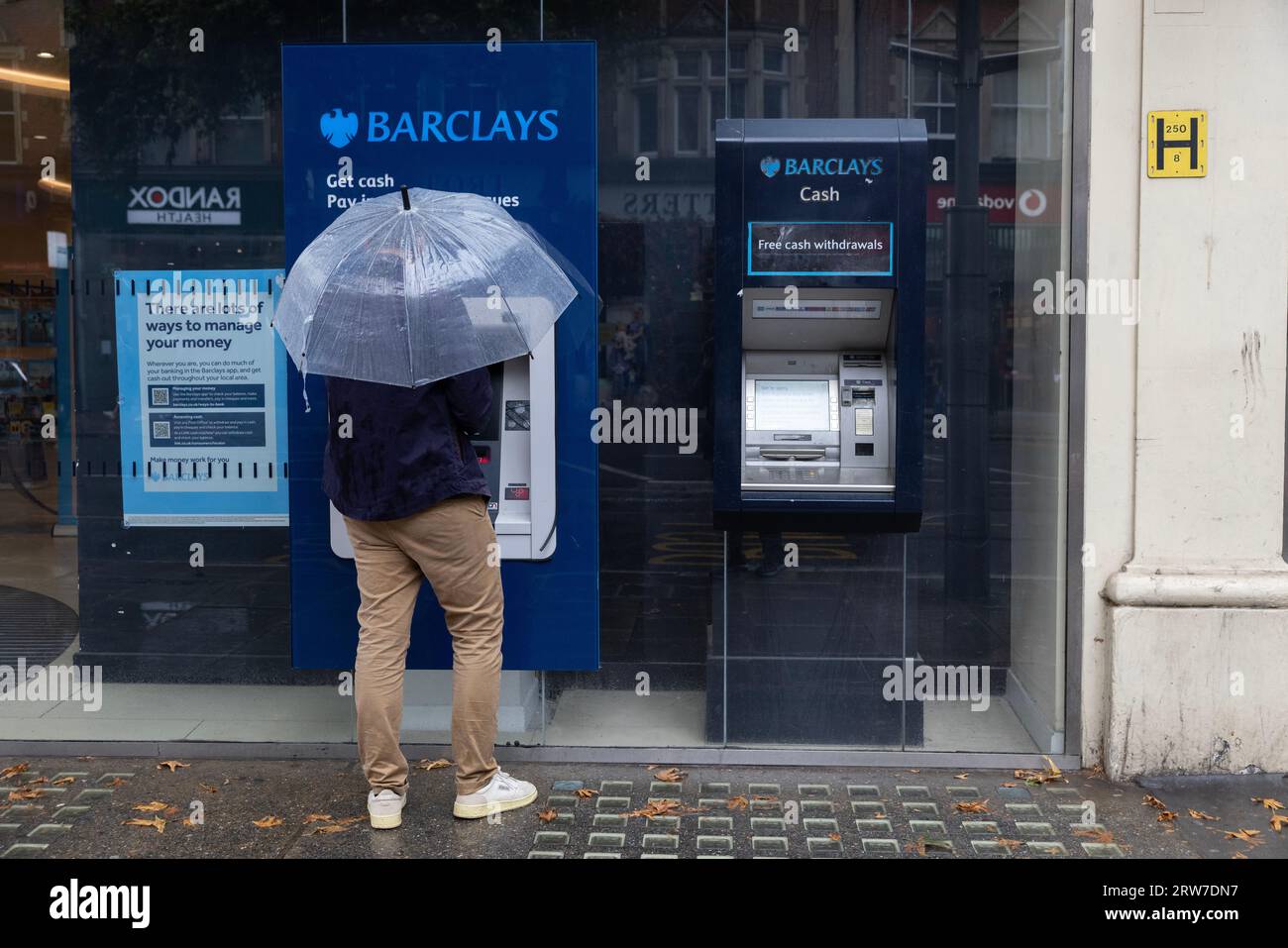 Uomo con ombrello che ritira contanti da un distributore di contanti della banca Barclays, High Street Kensington, Londra, Inghilterra, Regno Unito Foto Stock