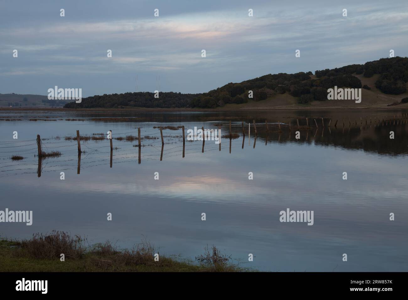 Una recinzione di legno e filo sta attraversando e un grande specchio d'acqua. L'acqua è blu. Ci sono delle colline dietro l'acqua. Il cielo è azzurro e nuvoloso Foto Stock