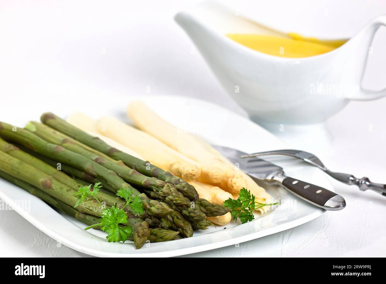 Gli asparagi verdi e bianchi giacciono su un piatto bianco su sfondo bianco. A destra, sullo sfondo, c'è una barca con salsa a canoa Foto Stock