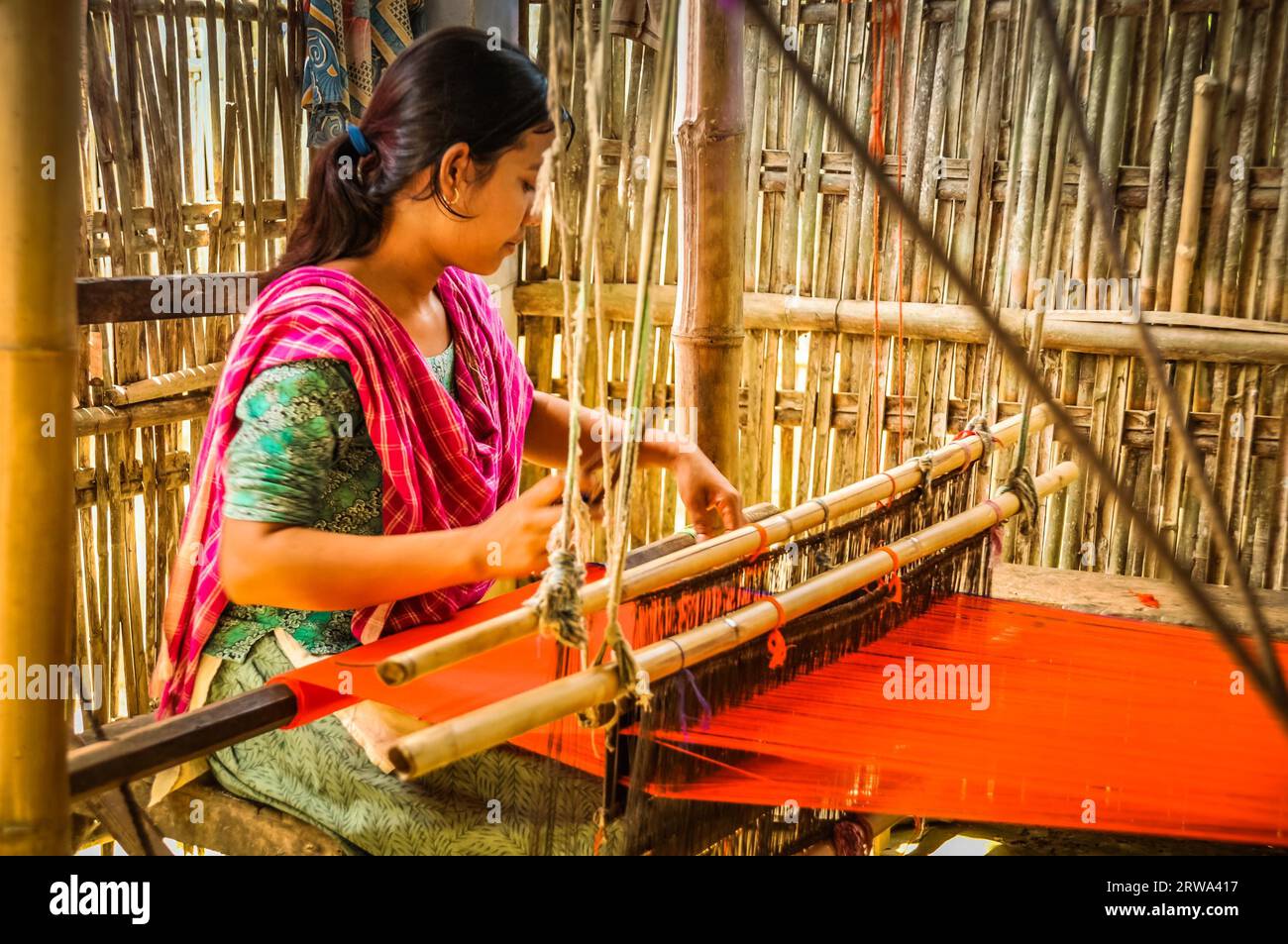 Srimongal, Bangladesh, circa luglio 2012: Giovane donna dai capelli neri con sciarpa rosa siede in un rifugio e si intreccia su un semplice telaio di legno a Srimongal Foto Stock
