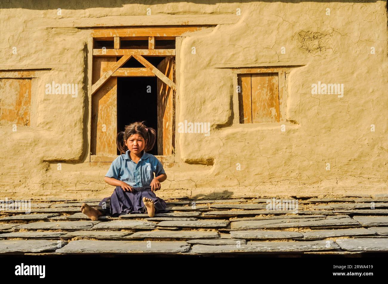 Beni, Nepal, circa maggio 2012: Una piccola ragazza in camicia blu e due code di cavallo siede a terra di fronte alla loro casa a beni, Nepal. Documentario editoriale Foto Stock