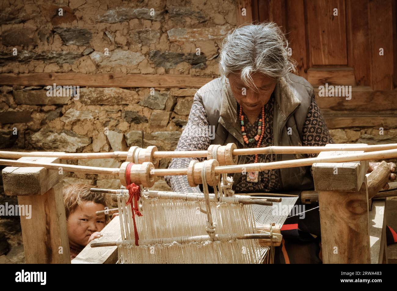 Dolpo, Nepal, circa giugno 2012: Vecchia donna dai capelli grigi vestita con gilet grigio con collane in perline guarda in basso e si intreccia su un semplice telaio di legno Foto Stock