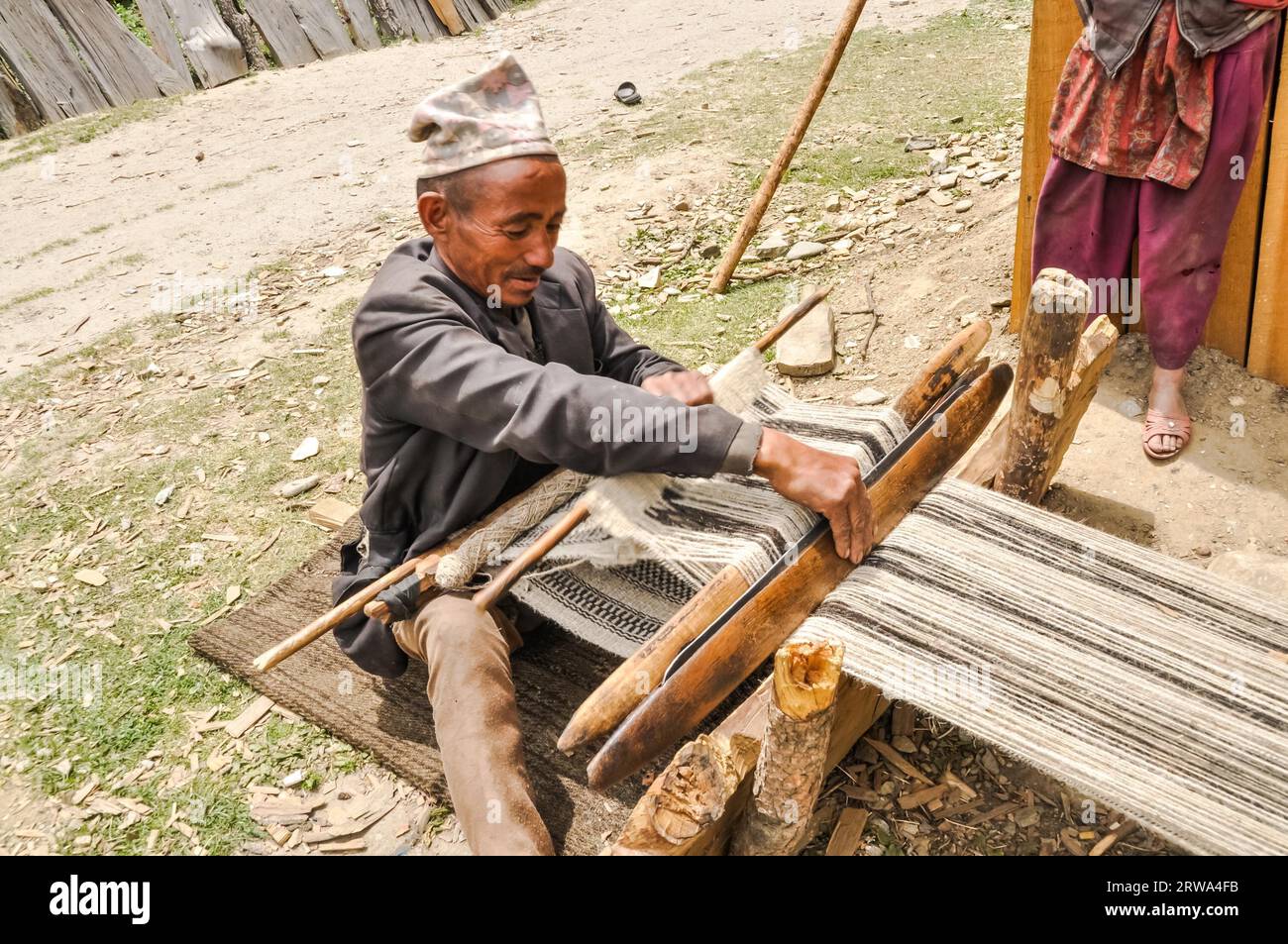 Dolpo, Nepal, circa giugno 2012: Uomo nativo in giacca grigia e berretto marrone intreccia su un semplice telaio per tessitura in legno e guarda in basso a Dolpo, Nepal. Foto Stock