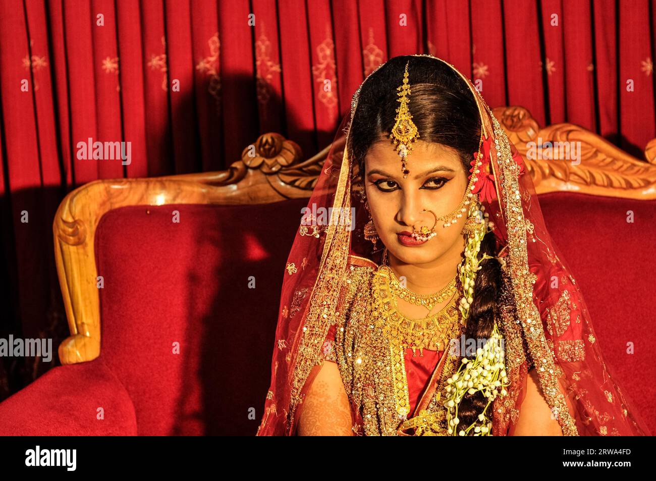 Dhaka, Bangladesh, circa luglio 2012: Giovane sposa dai capelli neri con un bellissimo abito rosso e oro scintillante siede sul divano rosso e sembra proprio a Dacca Foto Stock