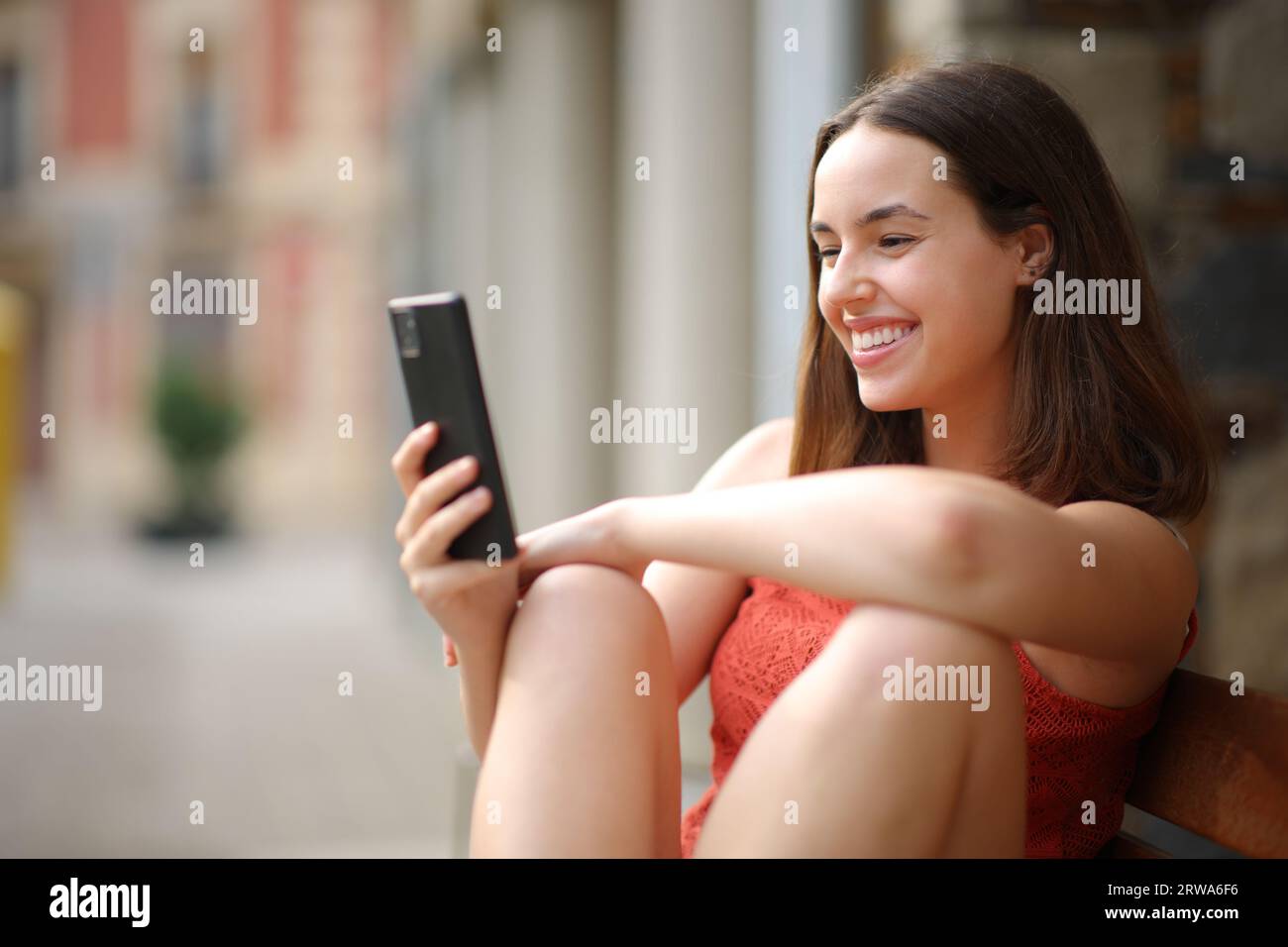 Donna felice seduta su una panchina in strada che usa uno smartphone Foto Stock