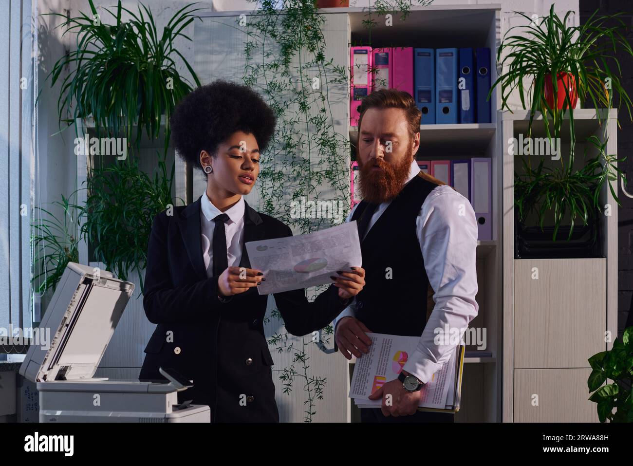 giovane segretaria afroamericana che mostra un documento a un uomo d'affari barbuto vicino alla copiatrice in carica Foto Stock
