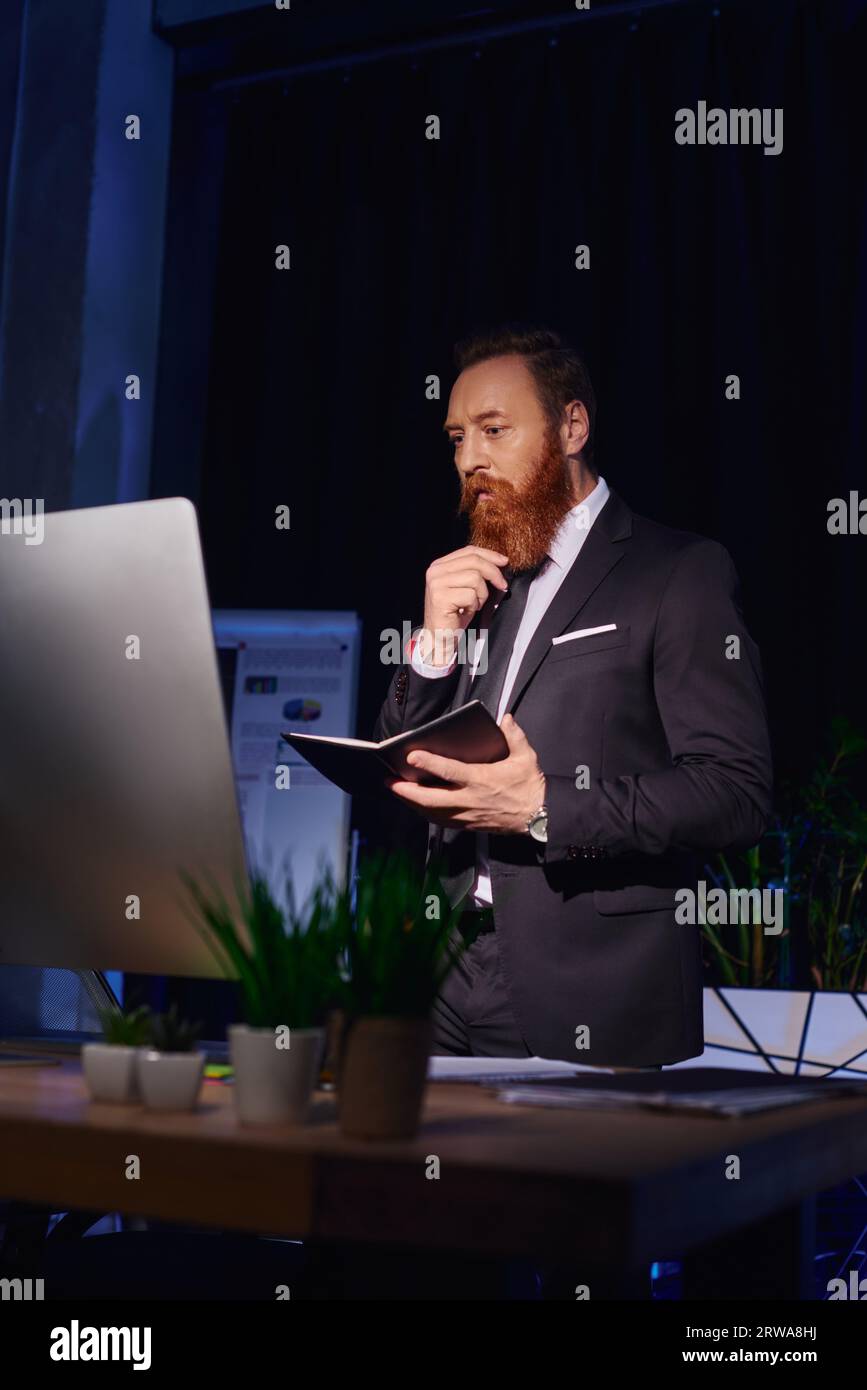 uomo d'affari con la barba premurosa con notebook che guarda il monitor del computer e lavora in ufficio notturno Foto Stock