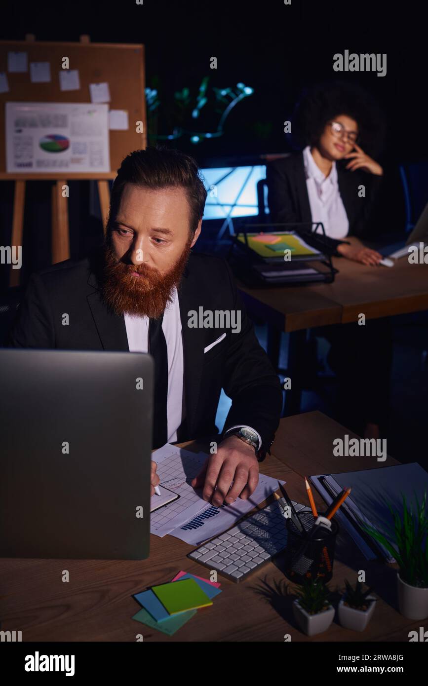 uomo barbuto che lavora con documenti e computer vicino all'affascinante donna afroamericana sullo sfondo Foto Stock