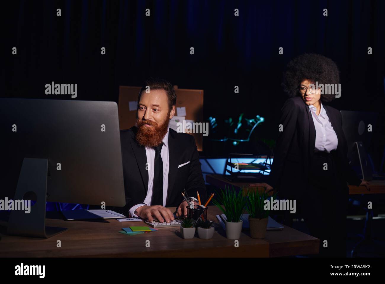 attraente donna afroamericana vicino a un uomo d'affari barbuto che lavora al computer di notte in ufficio Foto Stock