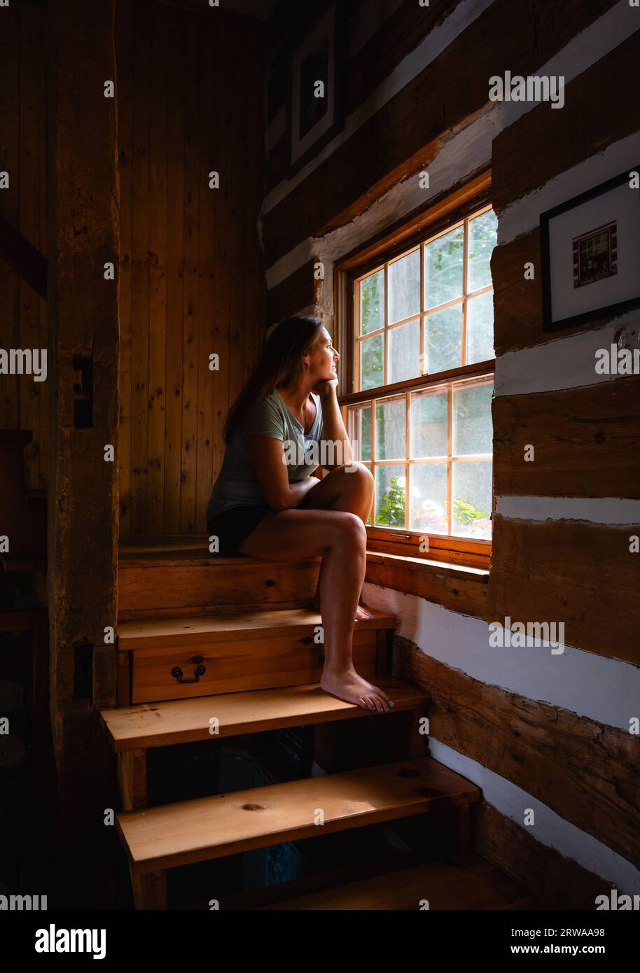 Donna seduta sulle scale della casa di legno che guarda fuori dalla finestra. Foto Stock