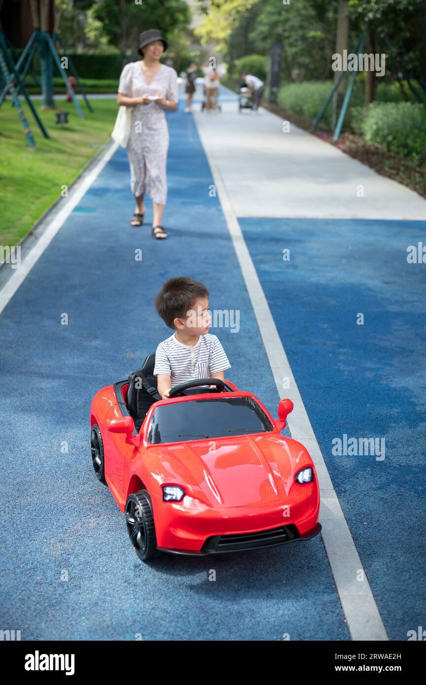 Un simpatico bambino multirazziale cavalca un'auto rossa con telecomando mentre sua madre cammina dietro di lui guidando un veicolo giocattolo Foto Stock