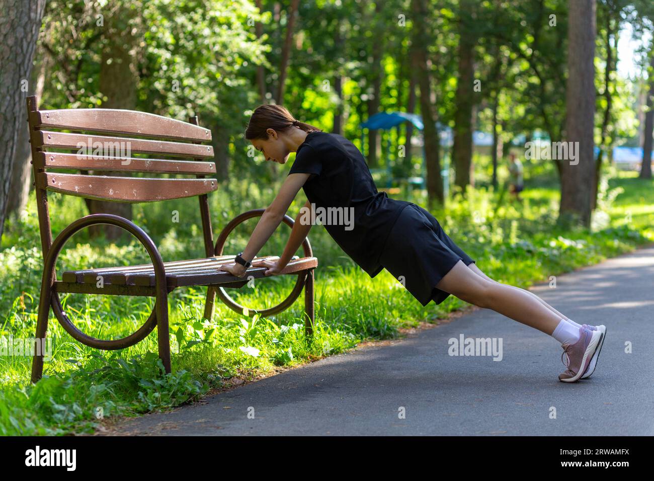 Bella giovane donna sportiva con t-shirt nera, pantaloncini neri e scarpe da ginnastica rosa che riscaldano i triceps e il torace facendo push-up dalla panchina AM Foto Stock