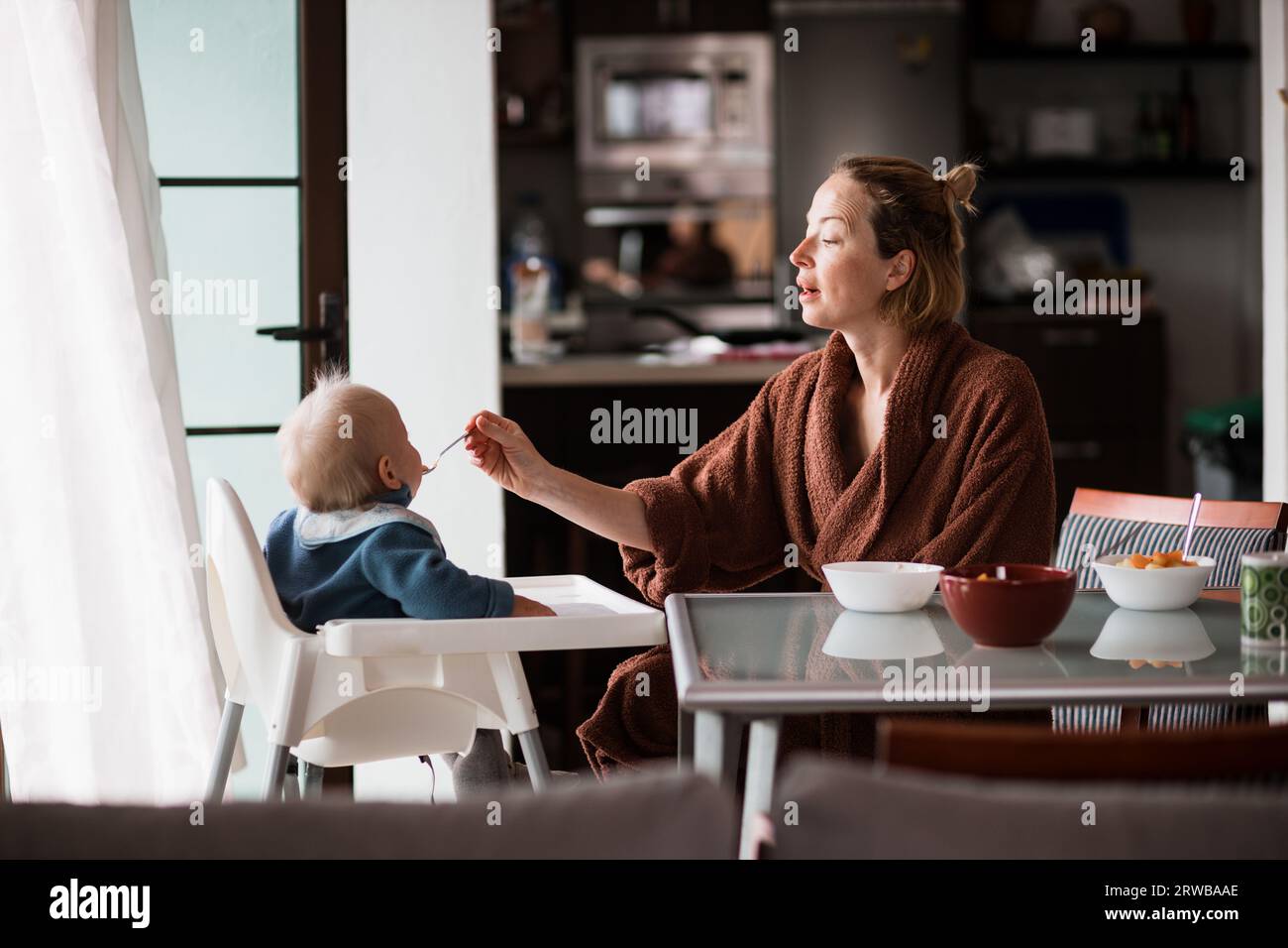 La mamma indossa il cucchiaio da bagno che allatta il bambino seduto in poltrona al tavolo da pranzo in cucina a casa la mattina. Foto Stock