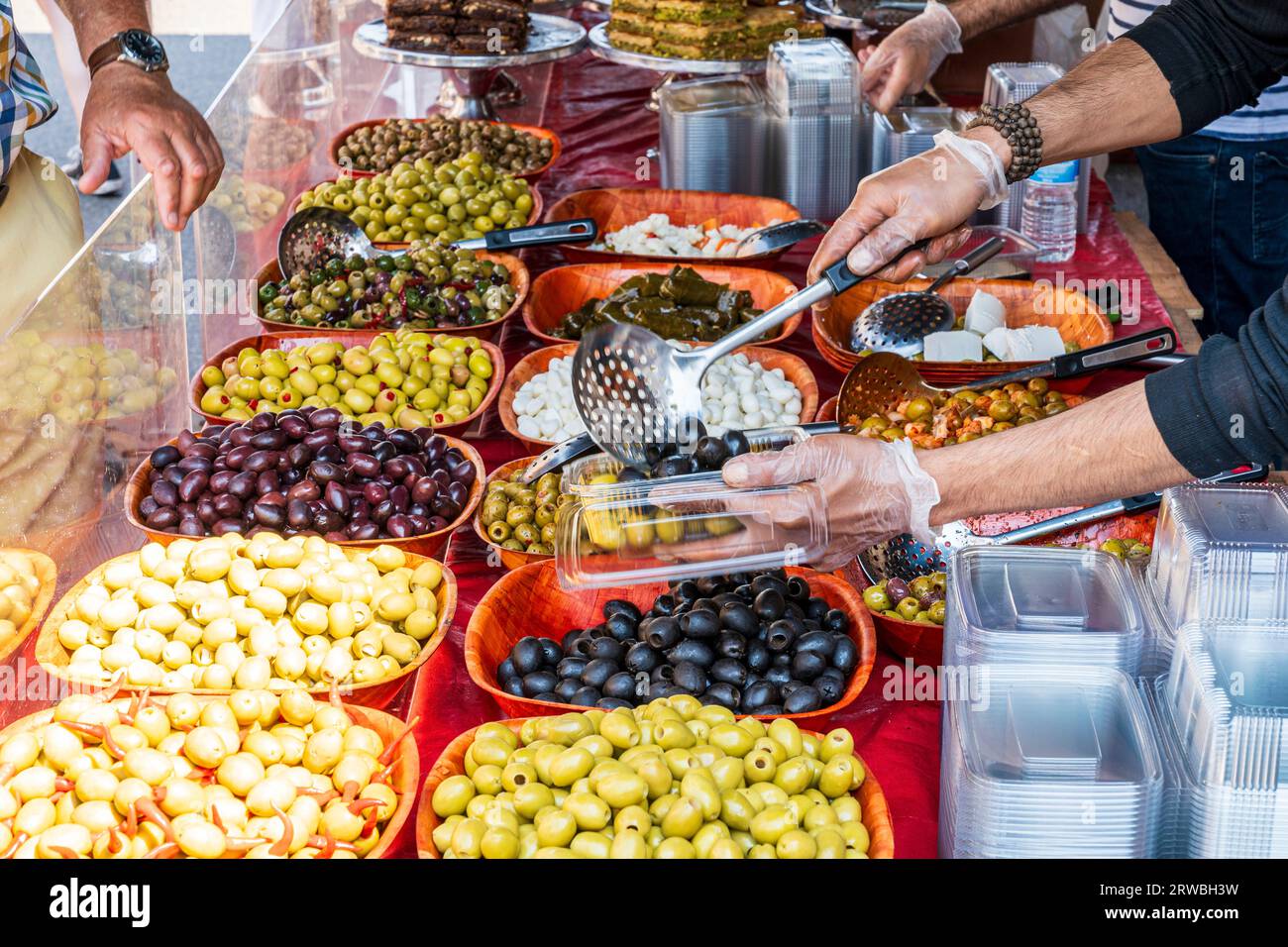 Primo piano di vari tipi di olive su una bancarella alimentare. Il braccio e le mani dell'uomo si avvicinano in avanti per raccogliere alcune olive in un contenitore di plastica da vendere. Foto Stock