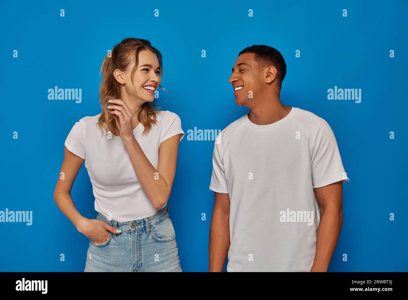 donna allegra che posa con la mano in tasca e guarda un amico afro-americano su sfondo blu Foto Stock