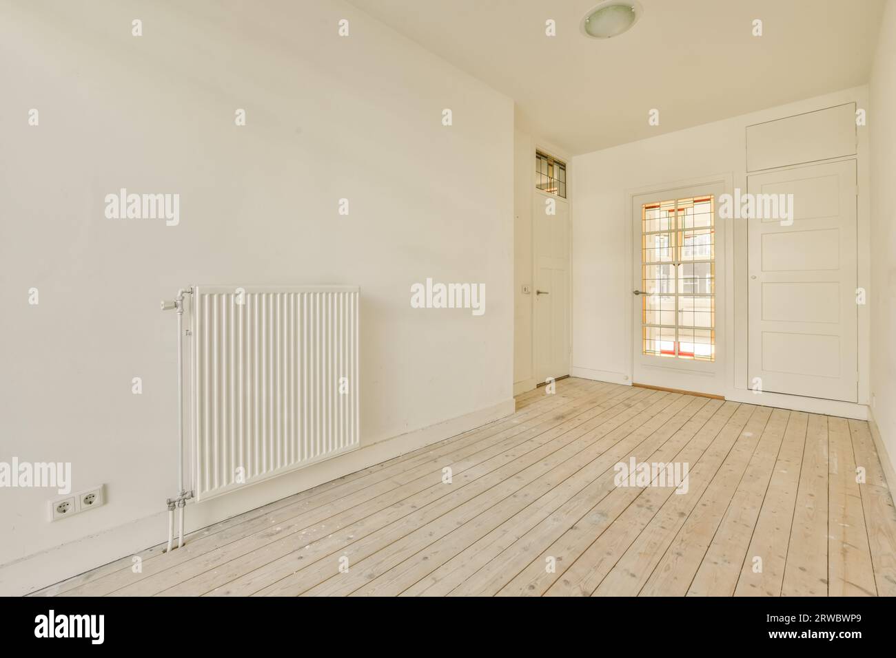 Interno di una spaziosa camera con radiatore a parete e porte chiuse in un moderno appartamento vuoto con pavimento in legno duro e pareti bianche Foto Stock