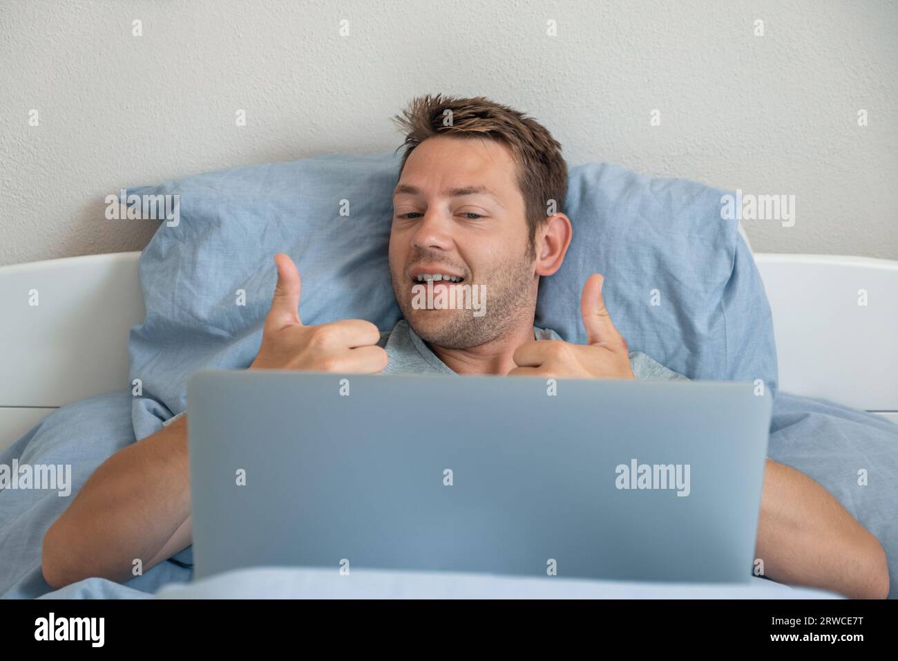 Bell'uomo caucasico sorridente e che mostra il gesto del pollice in alto durante la video chat sul laptop. Un uomo che si trova a letto e che ha una conversazione online la mattina Foto Stock