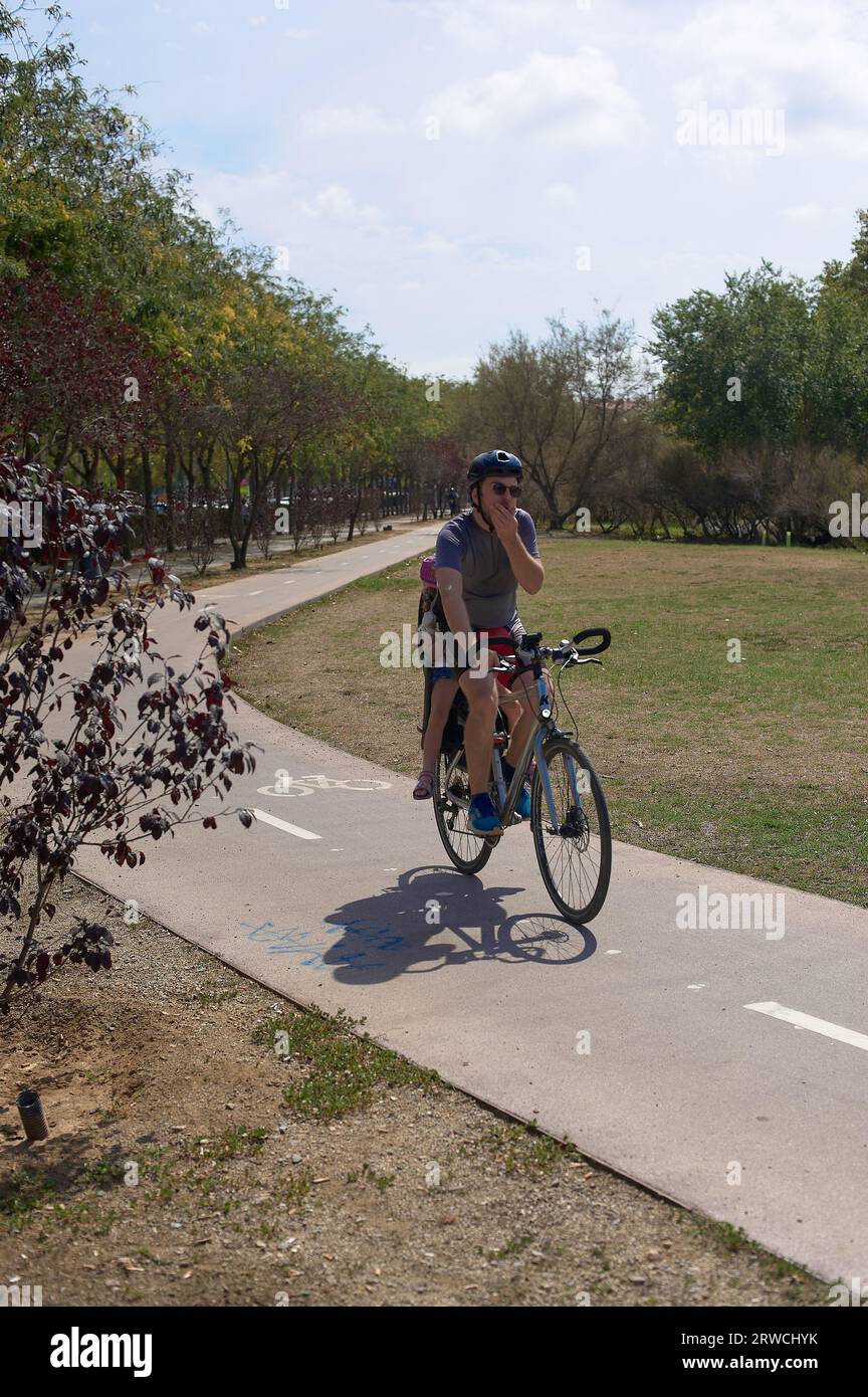 Viladecans, Spagna - 18 settembre 2023: Padre ciclista con sua figlia irriconoscibile seduta dietro sulla pista ciclabile nel mezzo di un parco pubblico Foto Stock