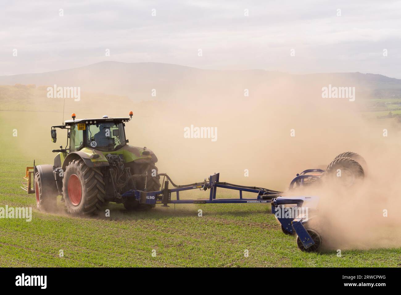 Un trattore Claas con rulli Dalbo che sfreccia nuvole di polvere in un campo asciutto di orzo primaverile Foto Stock