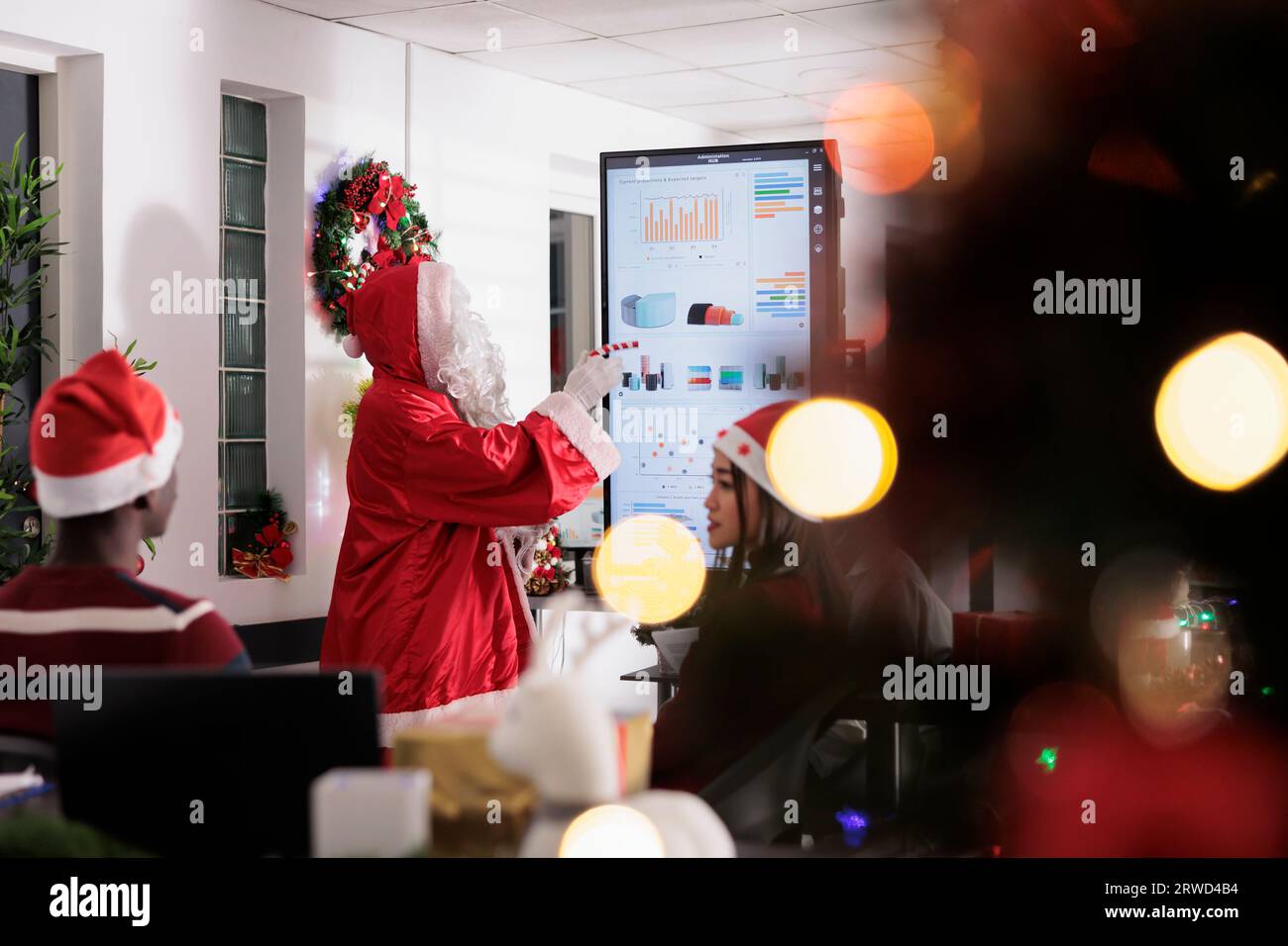 Leader del settore, vestito come Babbo Natale, offre ai dipendenti un'importante lezione nella sala riunioni ornata di Natale. Oratore aziendale che mostra i dati di visualizzazione delle statistiche aziendali ai lavoratori Foto Stock