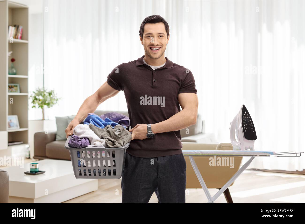 Un ragazzo che tiene un cestino pieno di vestiti davanti a un'asse da stiro in un soggiorno Foto Stock