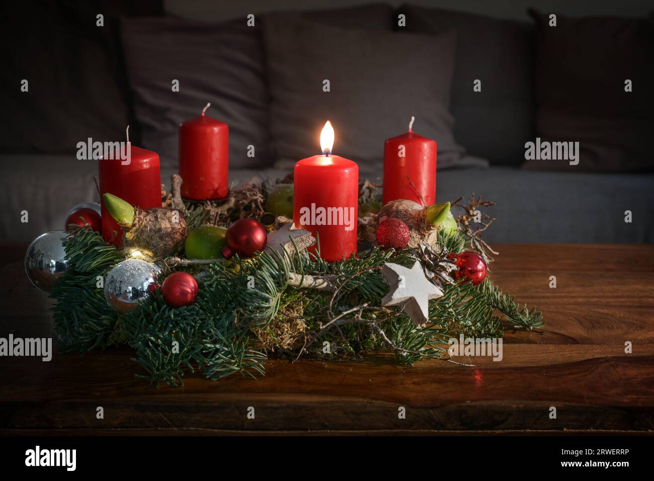 Corona dell'avvento con una candela rossa ardente e decorazione natalizia su un tavolo di legno davanti al divano, decorazioni casalinghe festive per la prima domenica, c Foto Stock