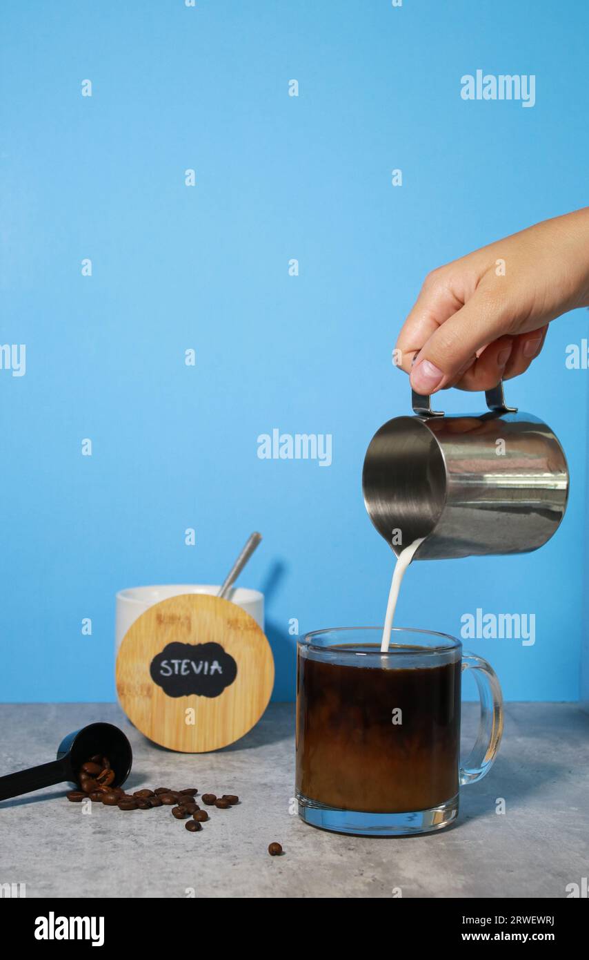 Versare il latte nel caffè nero in un bicchiere, stevia sullo sfondo Foto Stock
