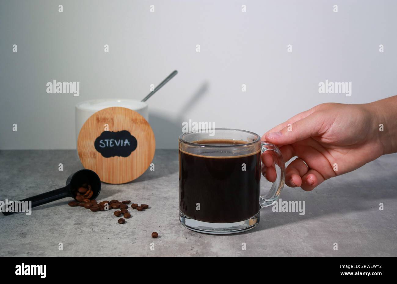 Mano che raggiunge un bicchiere di caffè nero e ciotola con dolcificante per stevia sullo sfondo Foto Stock