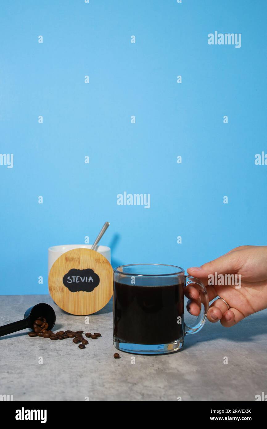 Mano che raggiunge un bicchiere di caffè nero e ciotola con dolcificante per stevia sullo sfondo Foto Stock