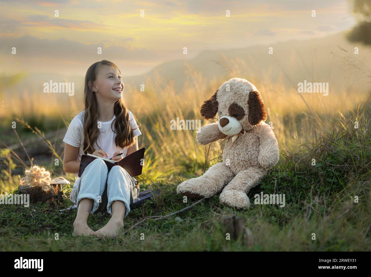 Ragazza giovane e sorridente seduta nella natura al tramonto con il suo orsacchiotto che scrive nel suo libro Foto Stock