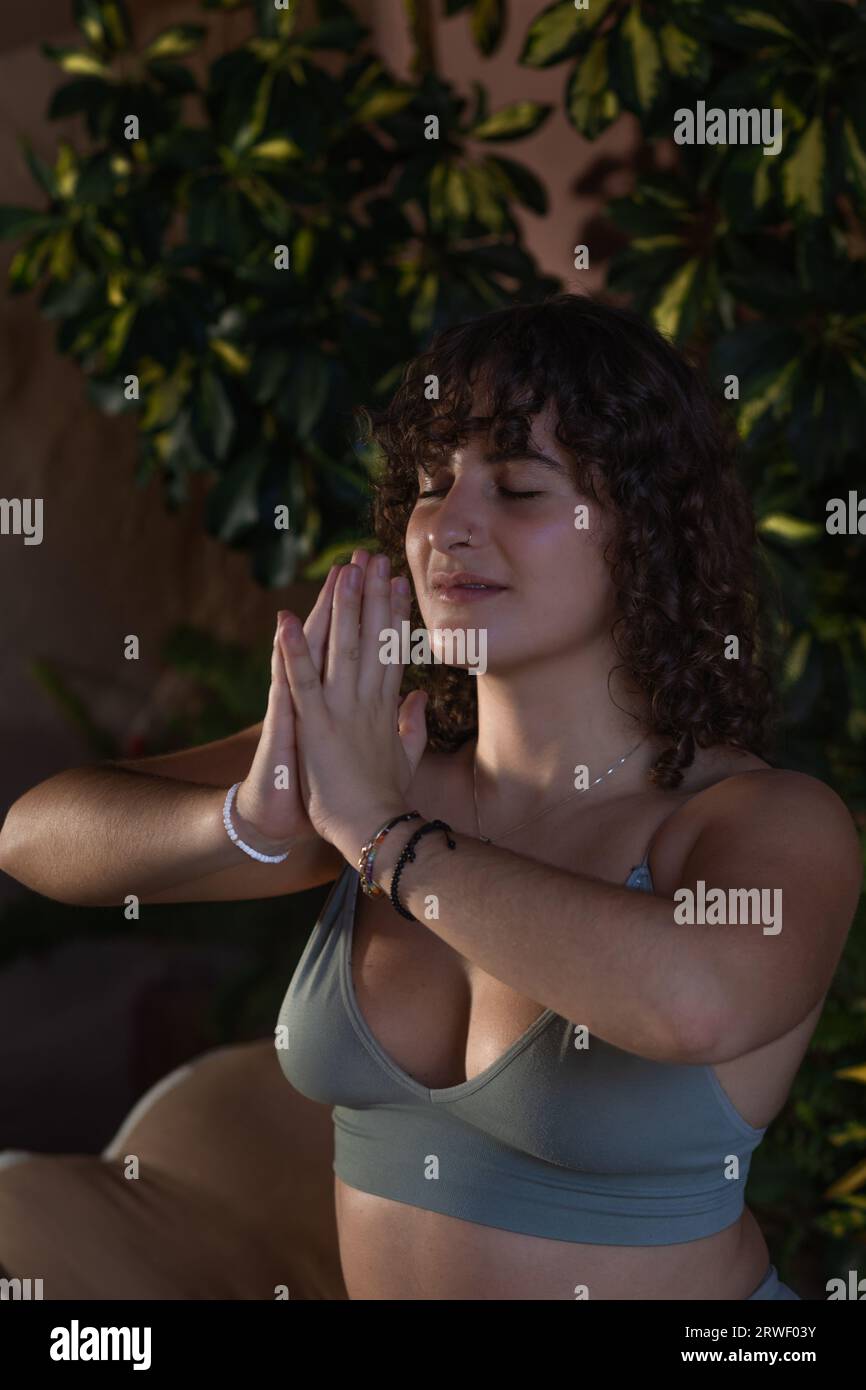 Ritratto ravvicinato di una giovane donna con capelli scuri ricci che meditano in un ambiente poco chiaro con le mani in posa di preghiera Foto Stock